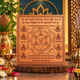 A square copper-colored spiritual plaque with engraved Sanskrit mantras and a central triangle diagram, displayed on a wooden stand in a decorative altar setting with candles, incense, flowers, and brass vessels.