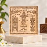 A square beige decorative plaque with engraved Hindu mantra and anatomical/mandala-style illustrations, displayed on a light marble pedestal in a softly lit indoor setting with blurred candles and flowers in the background.