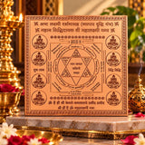 A square brass Vastu Purusha diagram plaque with Sanskrit mantras and illustrations, displayed on a marble altar surrounded by brass oil lamps, incense burner, and red rose petals in a temple setting.